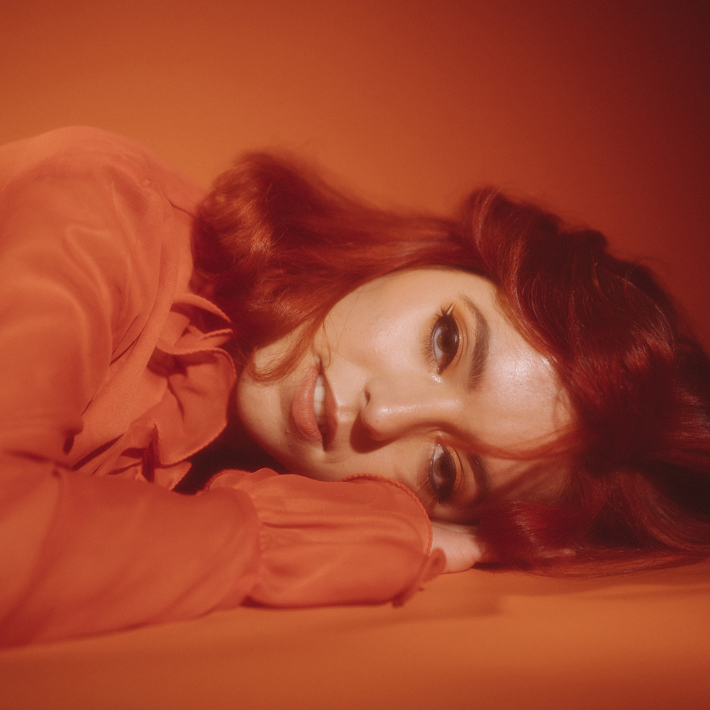 Emeree is laying on a red floor with their hand tucked under their head. They are wearing a bright red top and have long read hair.