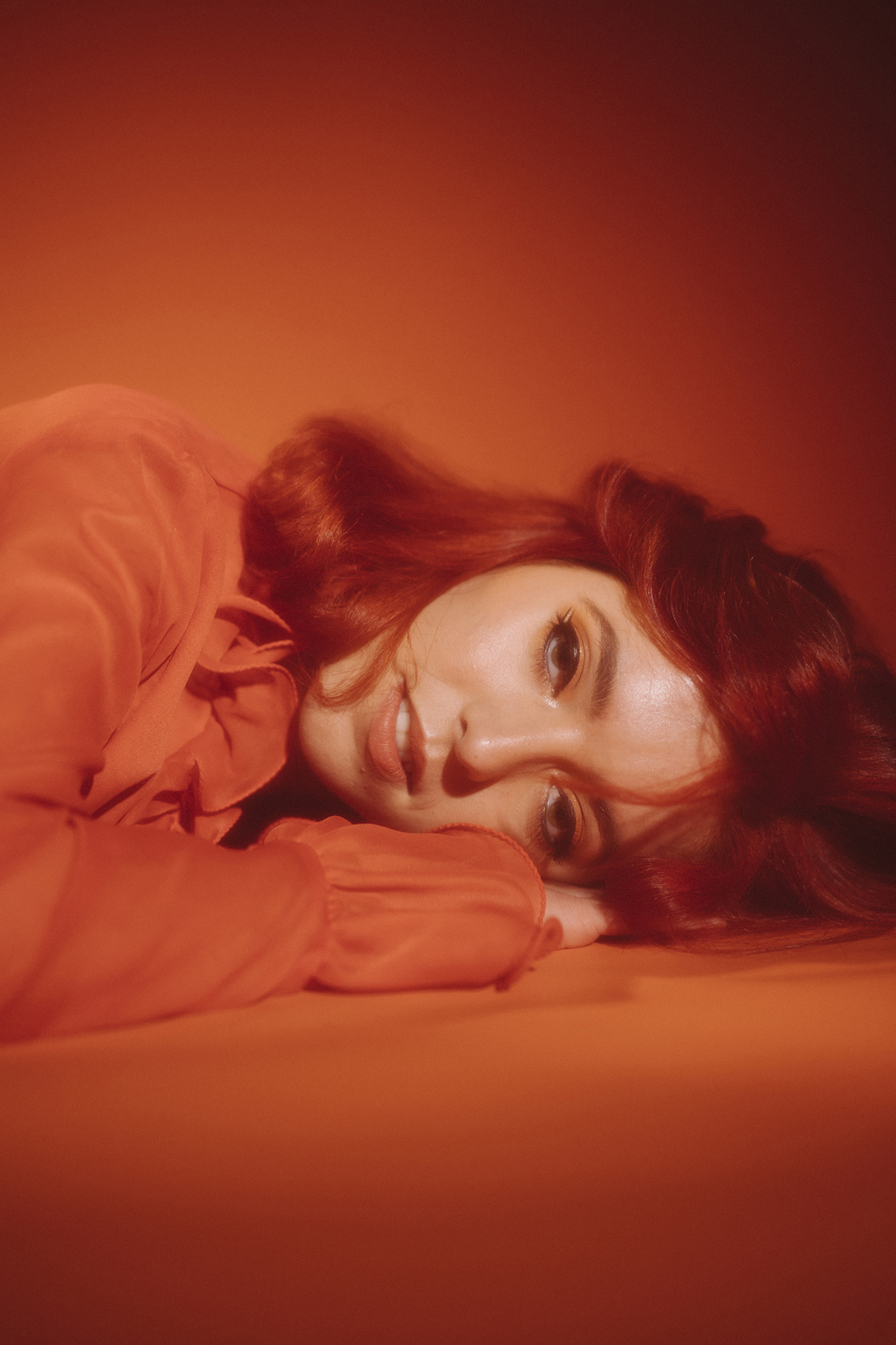 Emeree is laying on a red floor with their hand tucked under their head. They are wearing a bright red top and have long read hair.